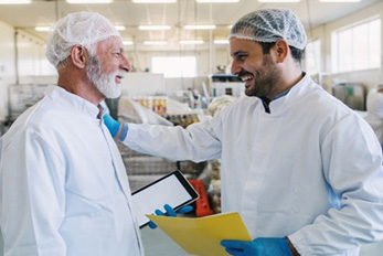 Two men in hair nets and white lab coats smiling at each other in food processing plant