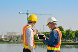 two men in hard hats and vests standing outside looking at tablet with crane in background