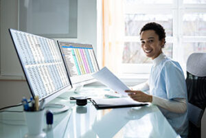 Woman in scrubs sitting at computer and smiling while working on medical billing and coding